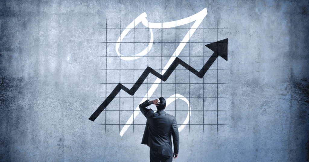 Image of a large graph with up-trending arrow and a % sign behind it, with a confused-looking man standing in front of it.